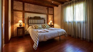 A bedroom with a brick wall, wooden beams, a bed with striped bedding, two bedside tables with lamps, and a large window with curtains.