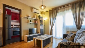 A modern living room with a sofa, a coffee table, a TV, and a kitchen area with red cabinets and a built-in oven. - AV Soterra a 3B in Vila (Ávila)