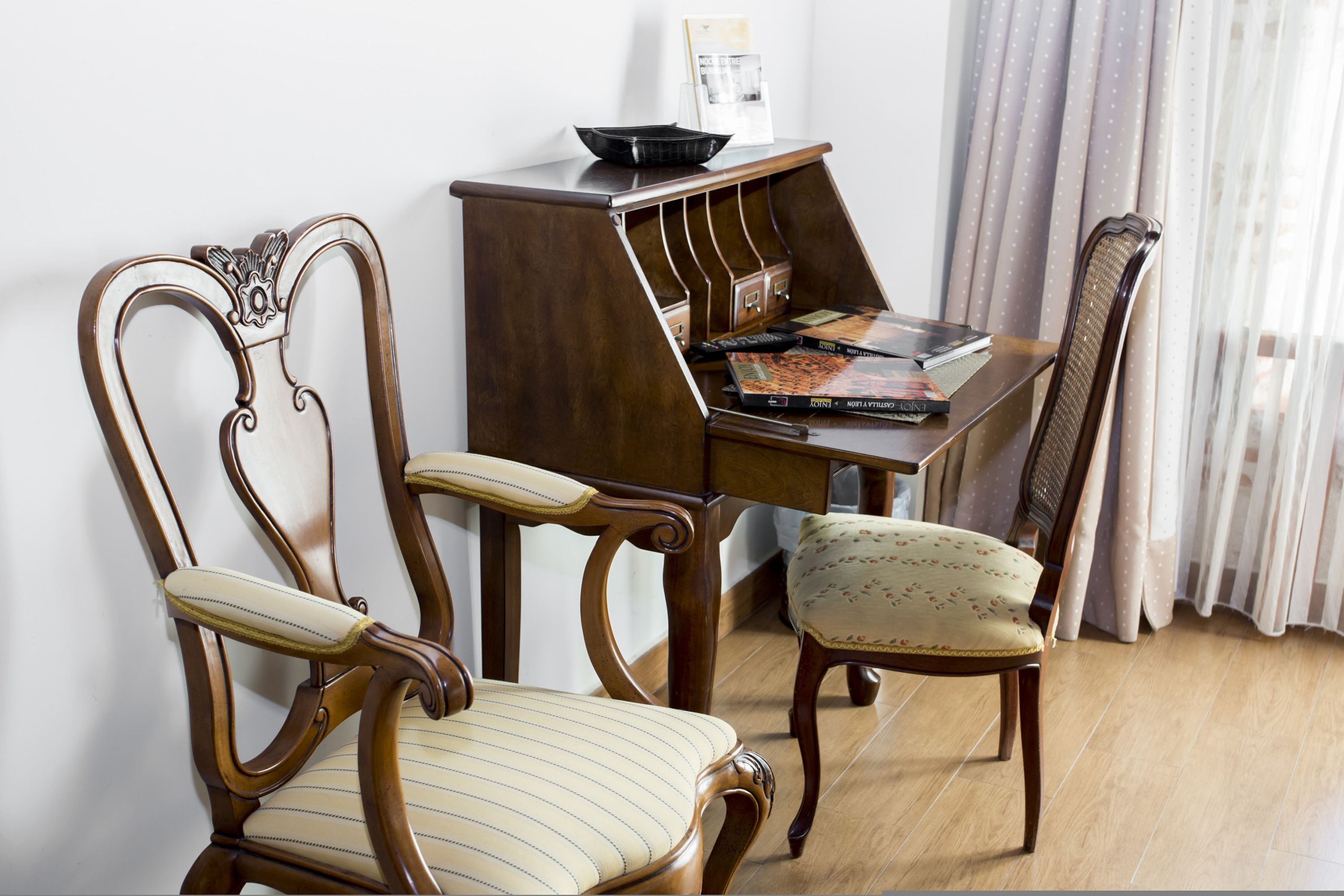 A room with two wooden chairs, one with a striped cushion and the other with a floral pattern. A wooden side table with a black bowl and a magazine is between the chairs.