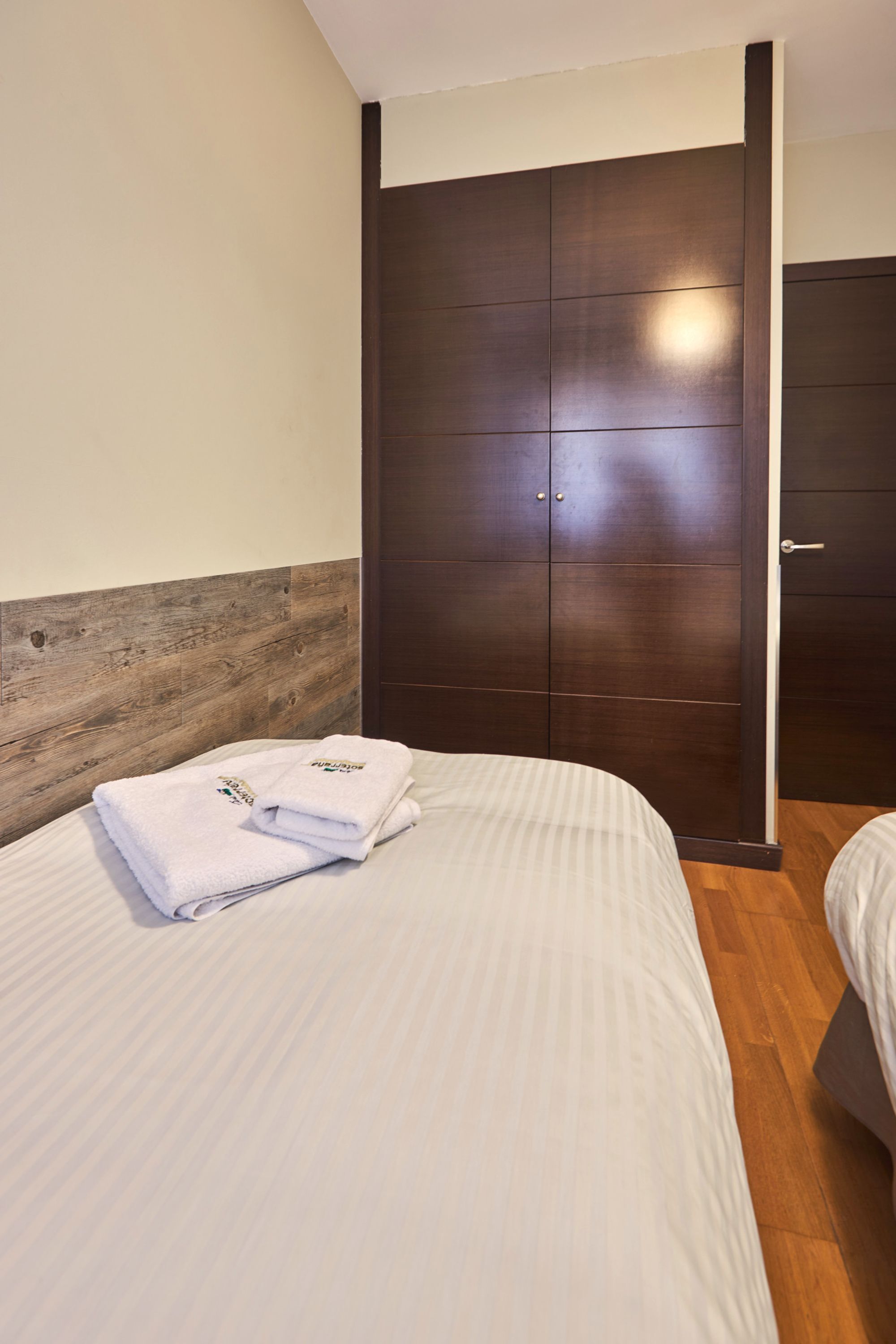 A hotel room with a wooden headboard, a large dark wooden wardrobe, and a neatly made bed with a folded white towel.