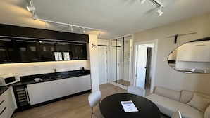 A modern kitchen with a black countertop, white cabinets, and a microwave. There is a dining area with a round table and four chairs. A sofa is visible in the background.