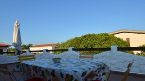 An outdoor dining area with a table set for two, covered with a tablecloth featuring a grapevine pattern. There are two chairs with red cushions, and a white umbrella is positioned to the left of the table. - Fpo-rose02p1 in Ottiolu (Budoni)