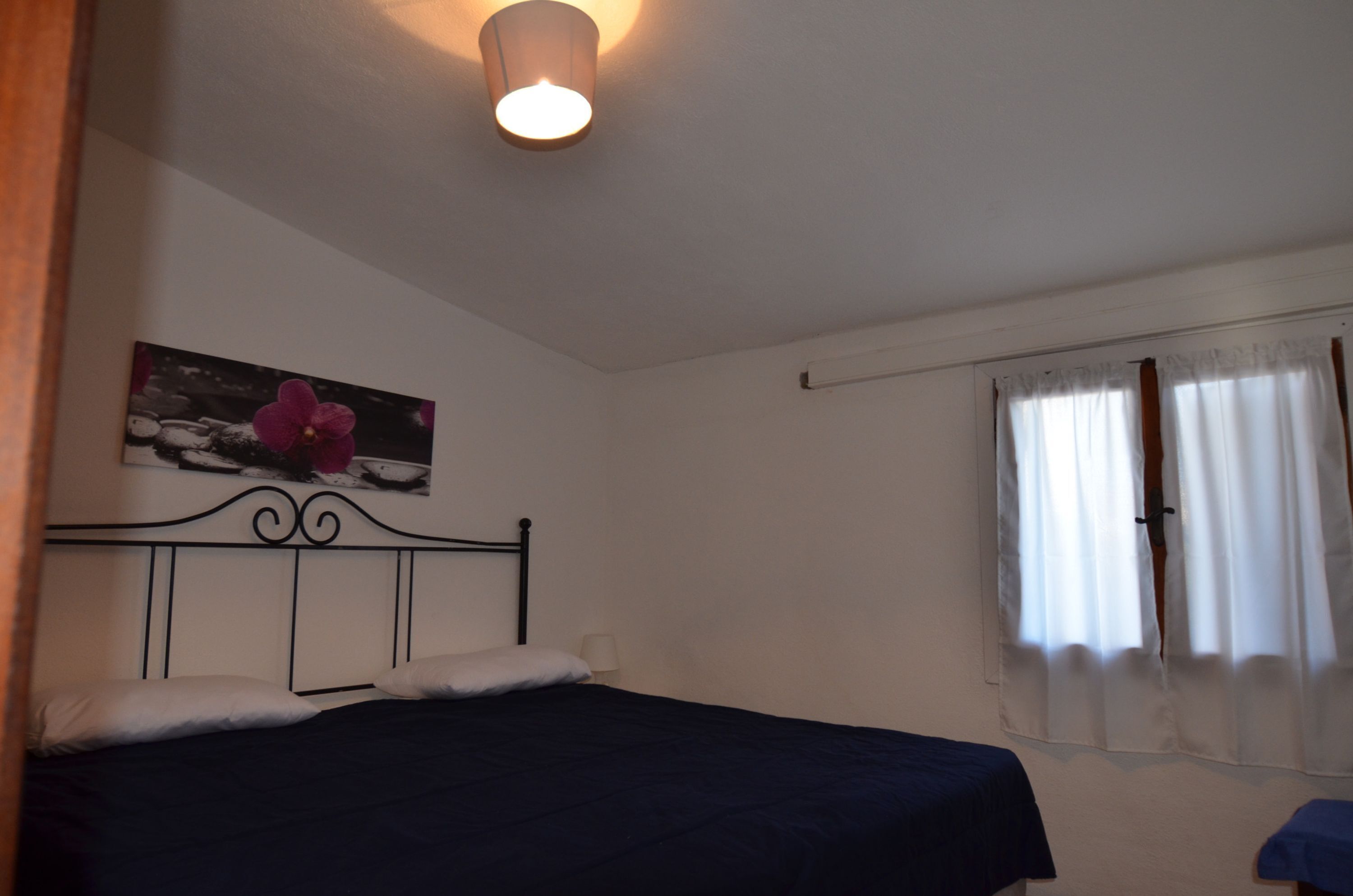Une chambre avec un lit simple, un luminaire fixé au mur, une fenêtre avec des rideaux et un tableau encadré accroché au mur.