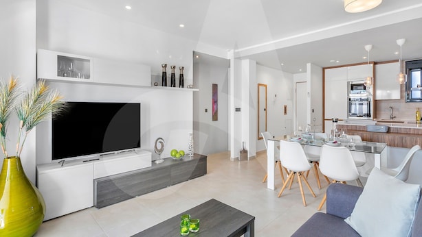 A modern living room with a flat-screen TV, a coffee table, and a sofa. There is a dining area with a table and chairs. The kitchen is visible in the background with modern appliances. - Casa Nicola in Guardamar (Guardamar del Segura)
