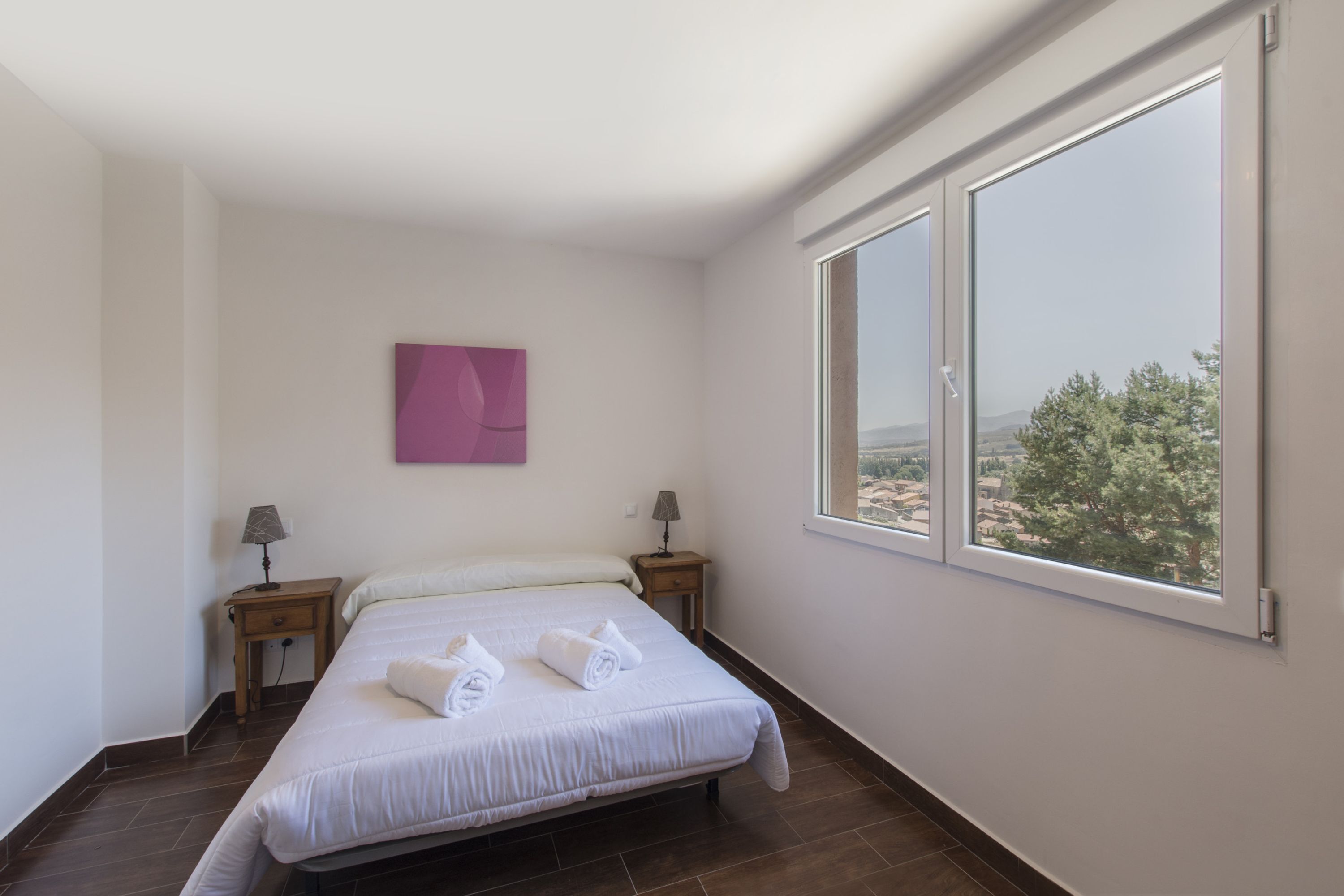 A bedroom with a large bed, two bedside tables, a window with a view, and a pink wall decoration.
