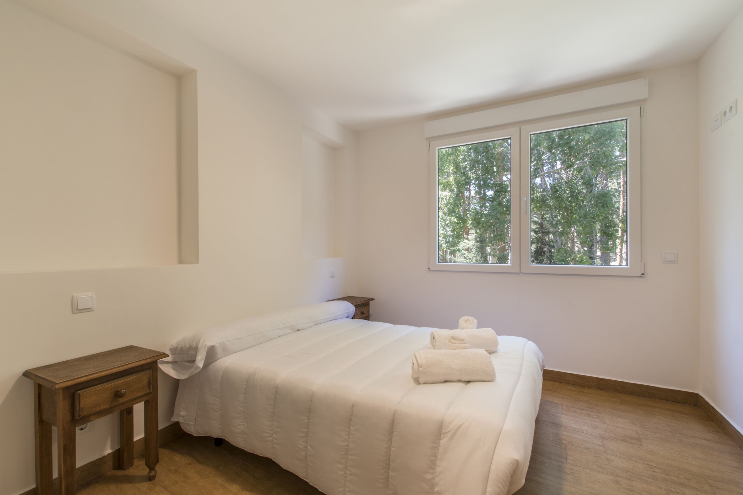 Ein Schlafzimmer mit Bett, Nachttisch, Fenster mit Blick auf Bäume und Lichtschalter an der Wand.