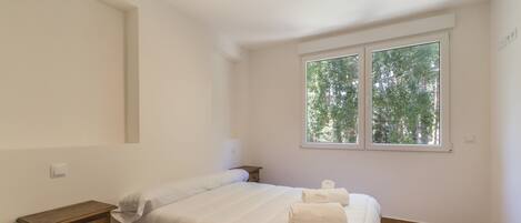A bedroom with a bed, a bedside table, a window with a view of trees, and a light switch on the wall.
