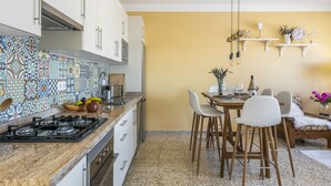 Een keuken met een betegelde spatwand, een gasfornuis, witte keukenkastjes en een eethoek met stoelen en een tafel.