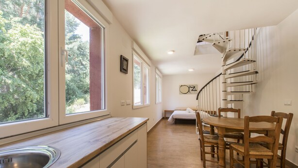 Une cuisine dotée d’un plan de travail en bois, d’un évier et d’un coin repas avec une table et des chaises en bois. Un escalier en colimaçon mène à un autre niveau.