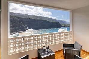 Ein Balkon mit Blick auf eine Küstenlandschaft, darunter Klippen und das Meer. Zwei Korbstühle und ein kleiner Tisch mit einer Flasche und Gläsern darauf.
