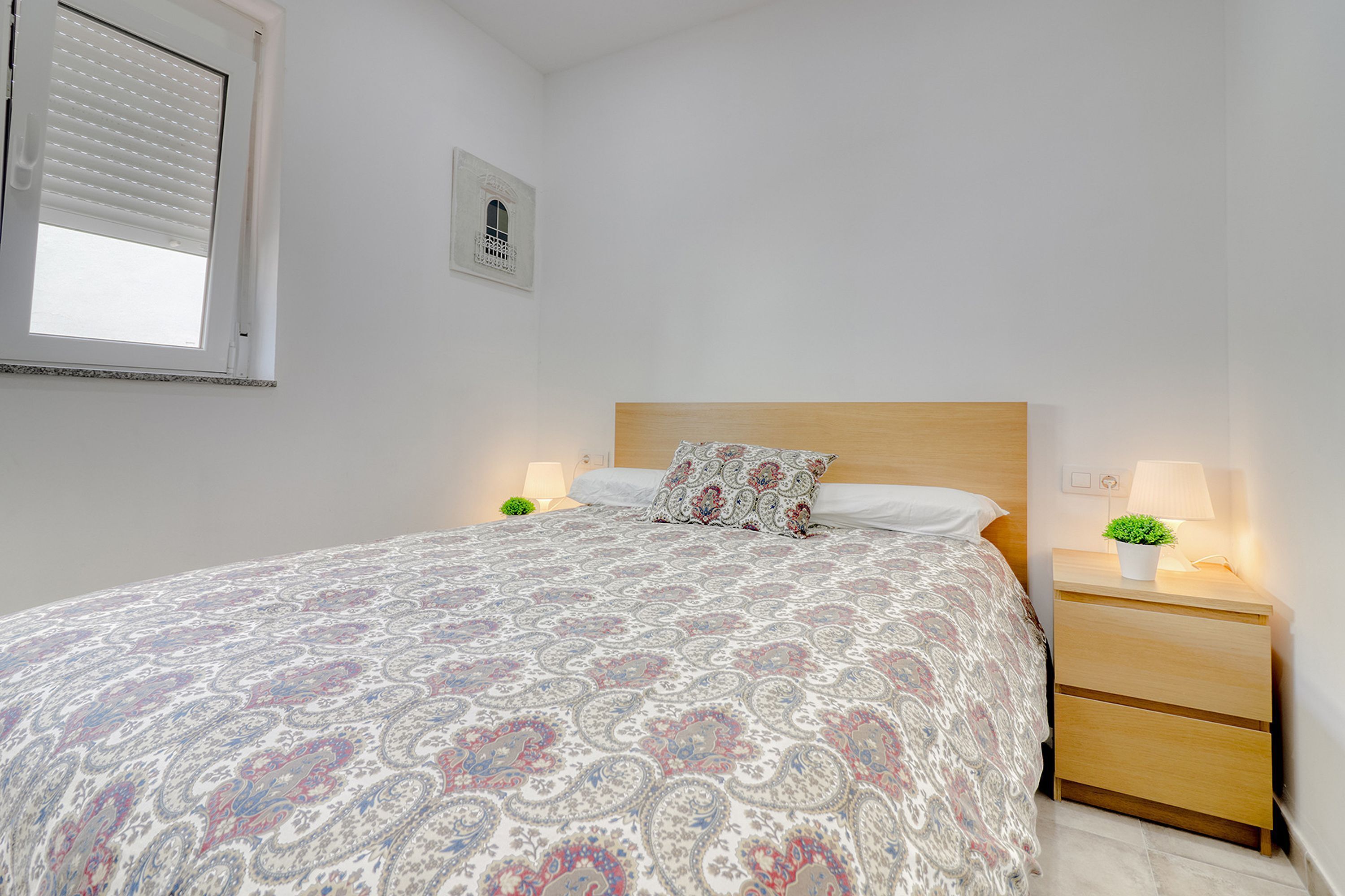 Une chambre avec un grand lit, deux tables de chevet avec des lampes, une fenêtre avec des stores et une petite décoration murale.