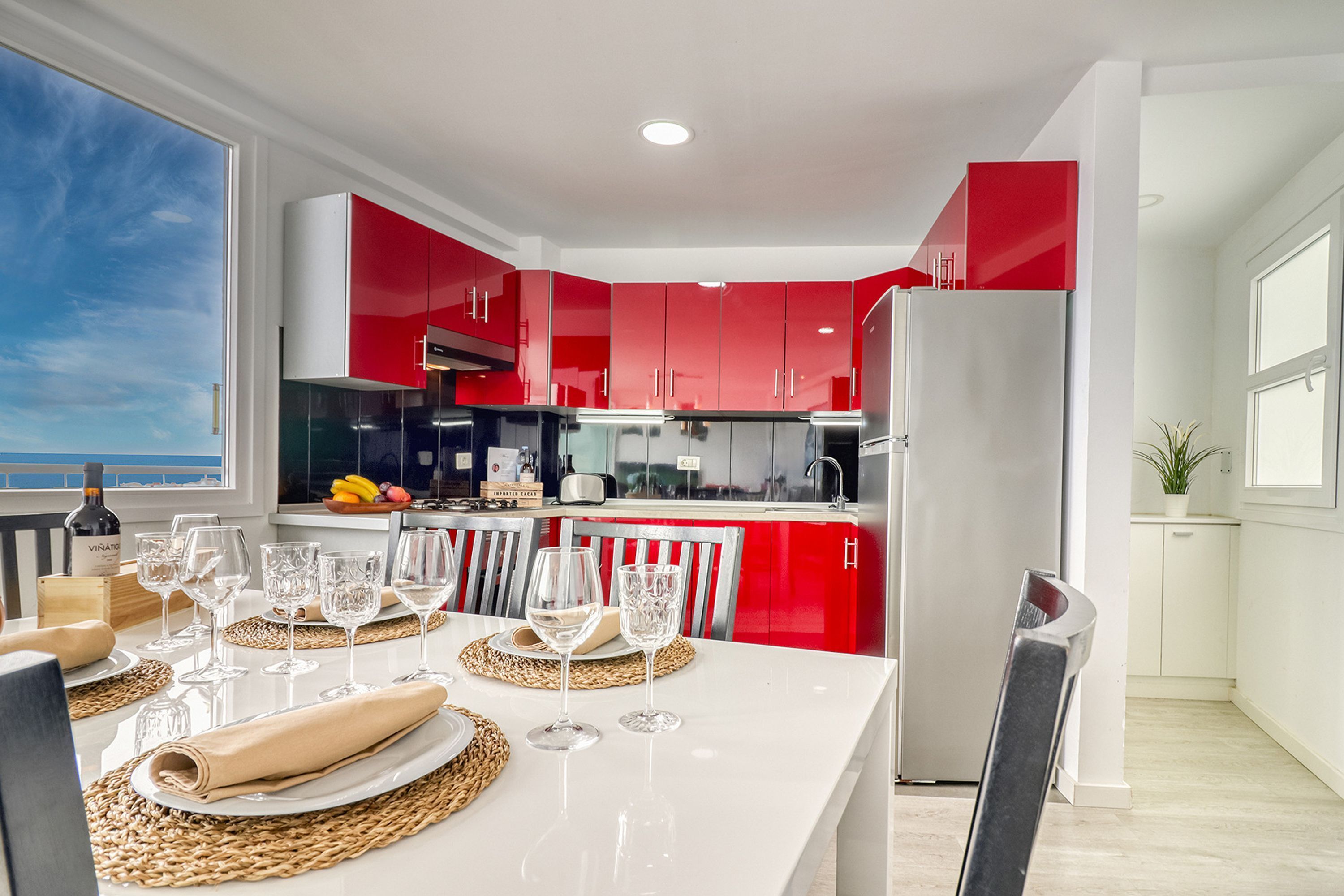 Une cuisine moderne avec des meubles rouges, un réfrigérateur blanc et un coin repas avec une table mise pour deux personnes.