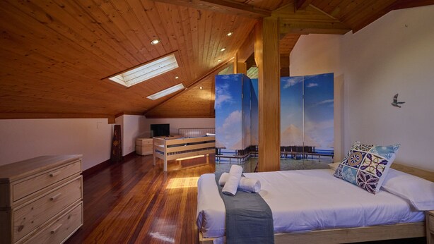 A bedroom with a wooden ceiling, skylight, a bed with white linens, a nightstand, a dresser, and a painting of a beach scene. - Great Escape by Fidalsa (Gautegiz Arteaga)