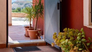 Puerta abierta que da a un balcón con plantas en macetas y vistas a zonas verdes y colinas.