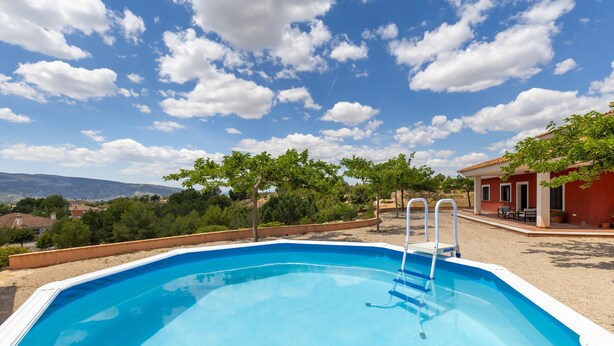 Una piscina rectangular con borde blanco, rodeada por una zona de arena y un edificio rojo con techo de tejas.