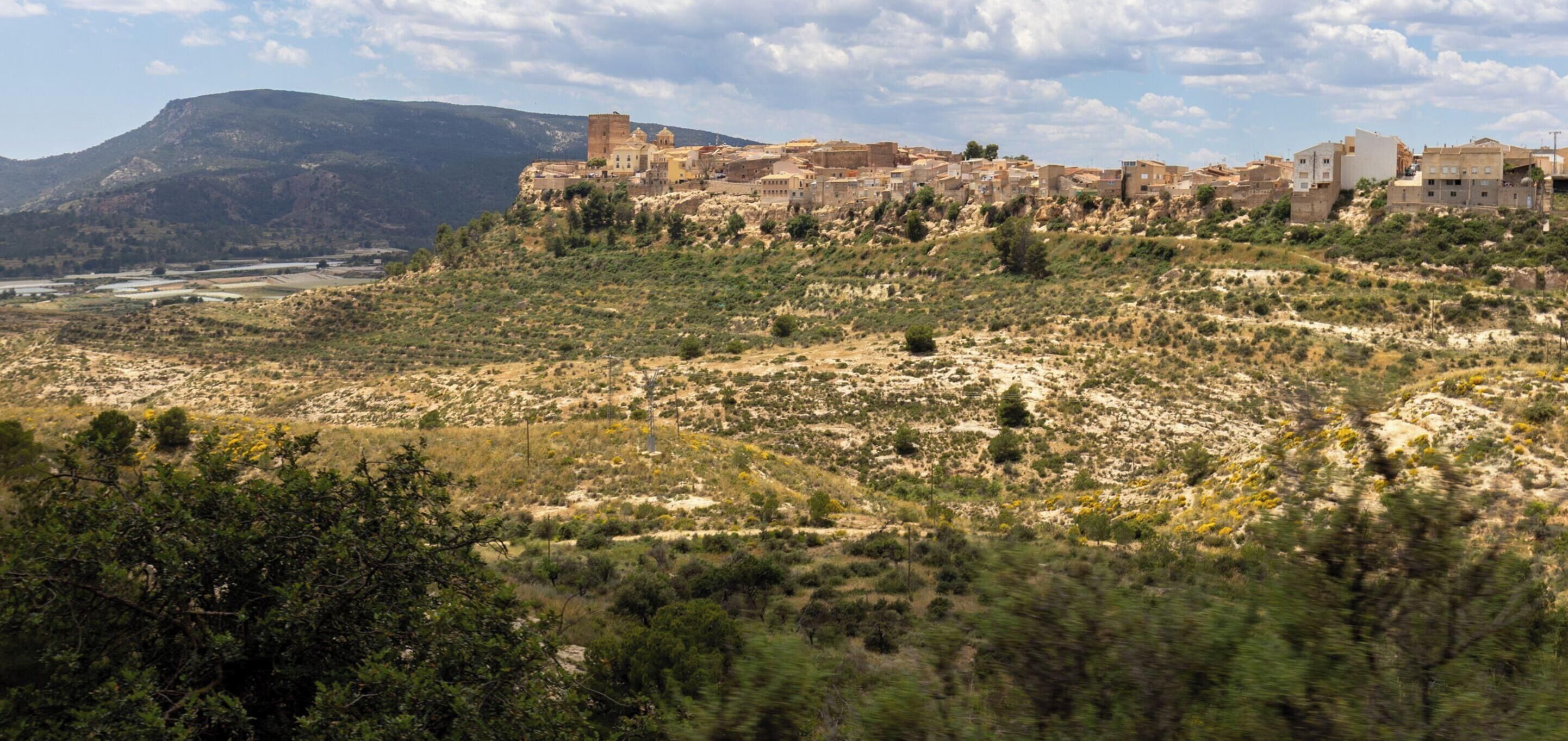 Un poble històric amb edificis antics en un turó, sota un cel clar, i la paraula "ALEDO" superposada.