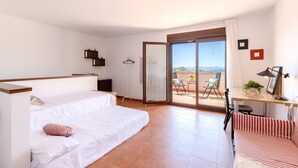 Un dormitorio moderno con cama, sofá, escritorio con lámpara y puerta corrediza de vidrio que da a un espacio exterior con mesa y sillas.