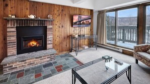 A living room with a fireplace, a television mounted on the wall, a glass coffee table, and a view of a snowy landscape through large windows.