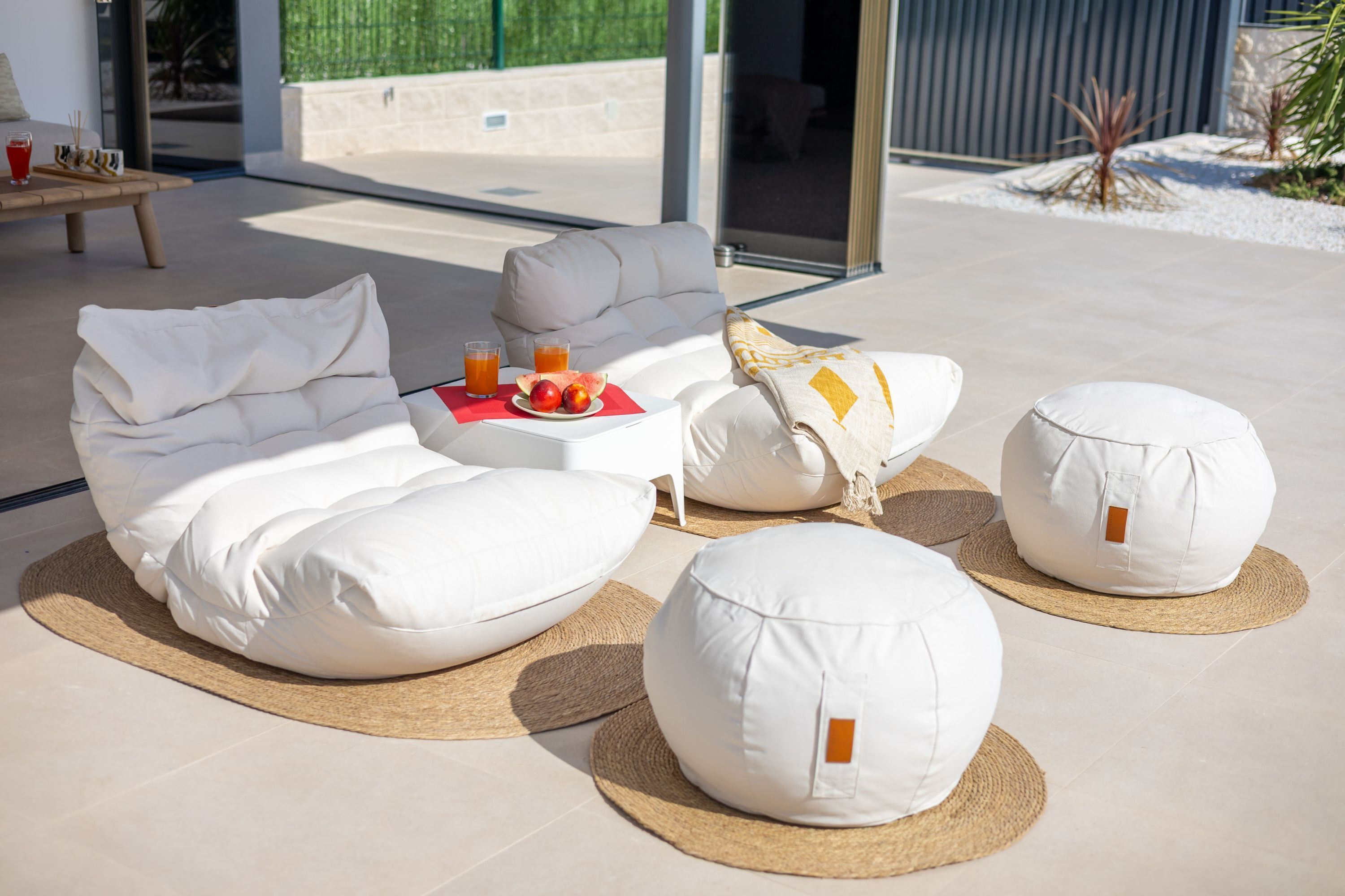 A modern outdoor seating area with a sofa, two bean bags, and a small table with drinks and fruit.