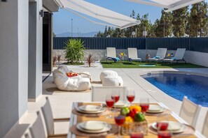 Uma área de refeições à beira da piscina, com uma mesa posta para dois, piscina e espreguiçadeiras.