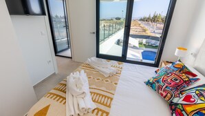 Une chambre d’hôtel avec un lit, un oreiller coloré, une serviette sur le lit, un téléviseur fixé au mur et une grande fenêtre donnant sur une piscine et un paysage.