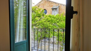 Una stanza con pavimento in legno, una porta scorrevole in vetro che dà su uno spazio esterno con un edificio in pietra, una recinzione metallica e un piccolo albero.