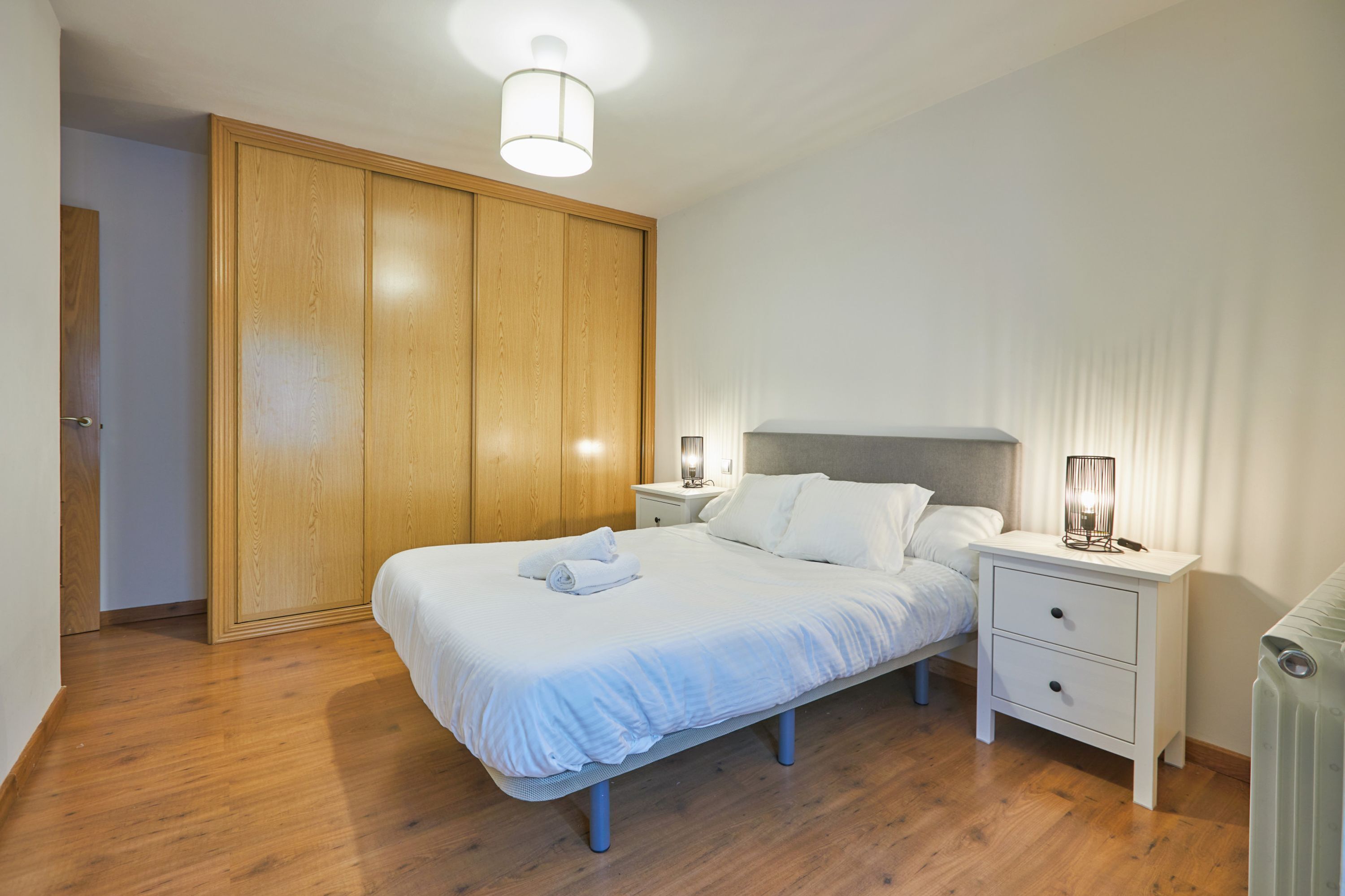 A bedroom with a wooden wardrobe, a bed with white bedding, a bedside table with a lamp, and a radiator.
