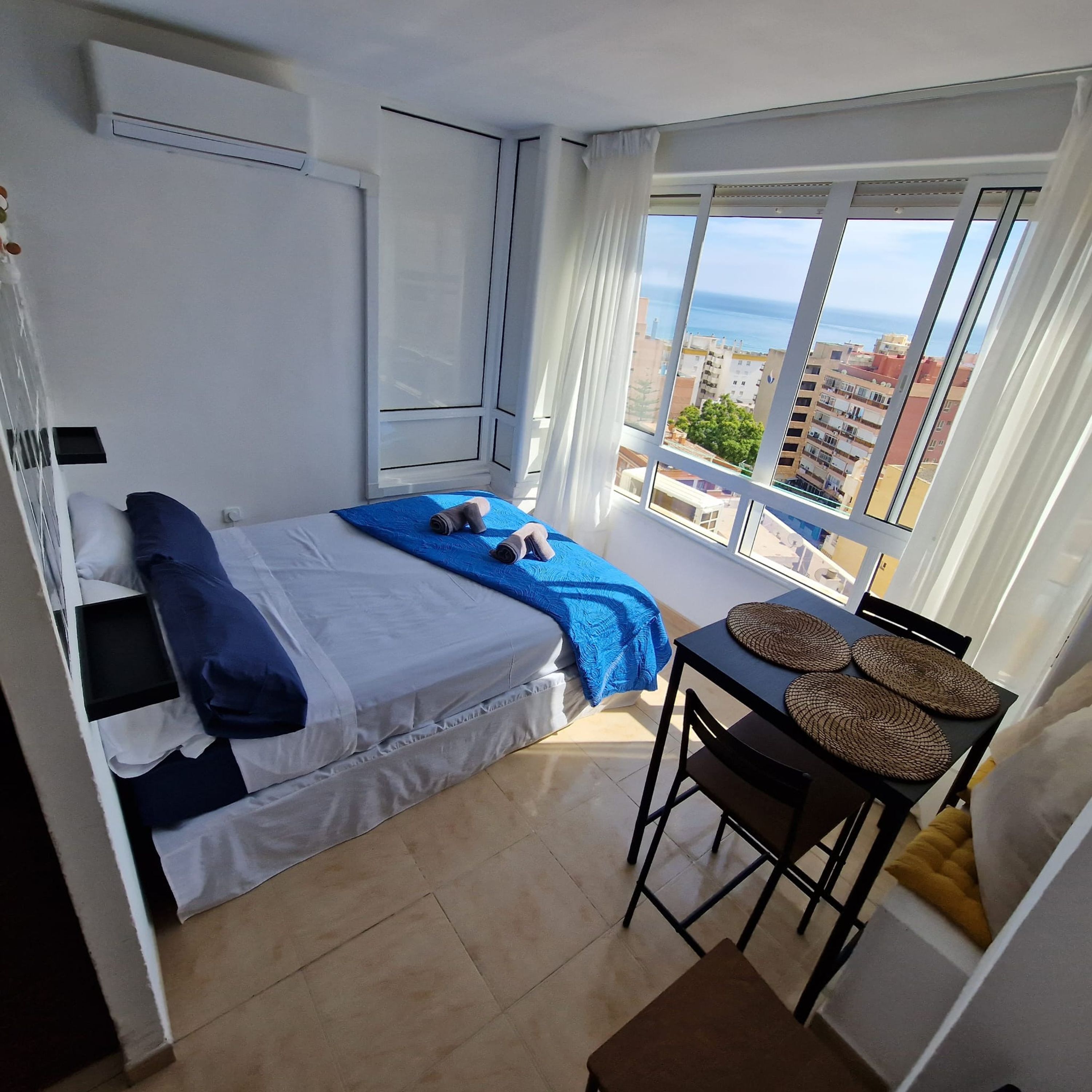 A bedroom with a bed, a small table with placemats, and a view of buildings and the sea through large windows.