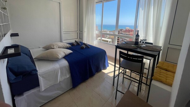A hotel room with a bed, a small table with a bottle and glass, a chair, and a view of the sea and buildings. - Tina de Steram Flats Torremolinos (Torremolinos)