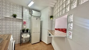 A compact kitchen with a refrigerator, a sink, and a countertop. There is a small shelf with plants and a red cushioned bench.