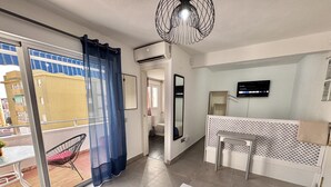 Una sala de estar moderna con una puerta corrediza de vidrio que da a un balcón, un televisor de pantalla plana montado en la pared y una pequeña mesa con una silla.