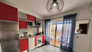 Una cocina compacta con armarios rojo y blanco, un refrigerador Bosch, un microondas y un lavamanos. Hay una mesa de comedor con sillas y un balcón con cortinas azules.
