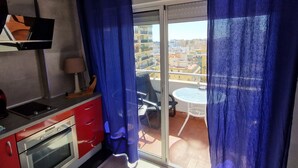 Un balcón con vistas a edificios, una puerta corredera de vidrio, una mesita y una zona de cocina roja con horno.