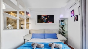 Una habitación de hotel moderna con una cama, ropa de cama azul y blanca, un cuadro grande en la pared y un armario de vidrio con plantas.