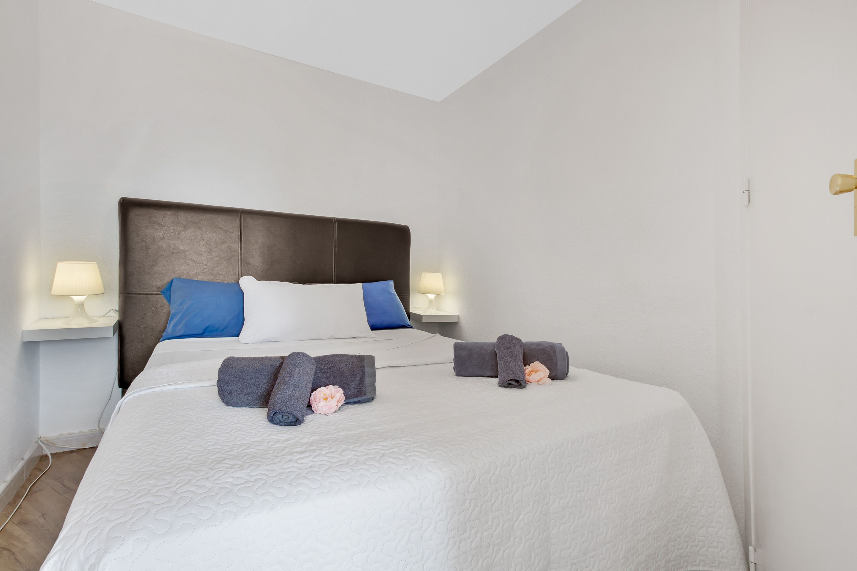 Una cama bien hecha con ropa de cama blanca, dos almohadas y toallas enrolladas. Hay lámparas de noche a ambos lados de la cama.