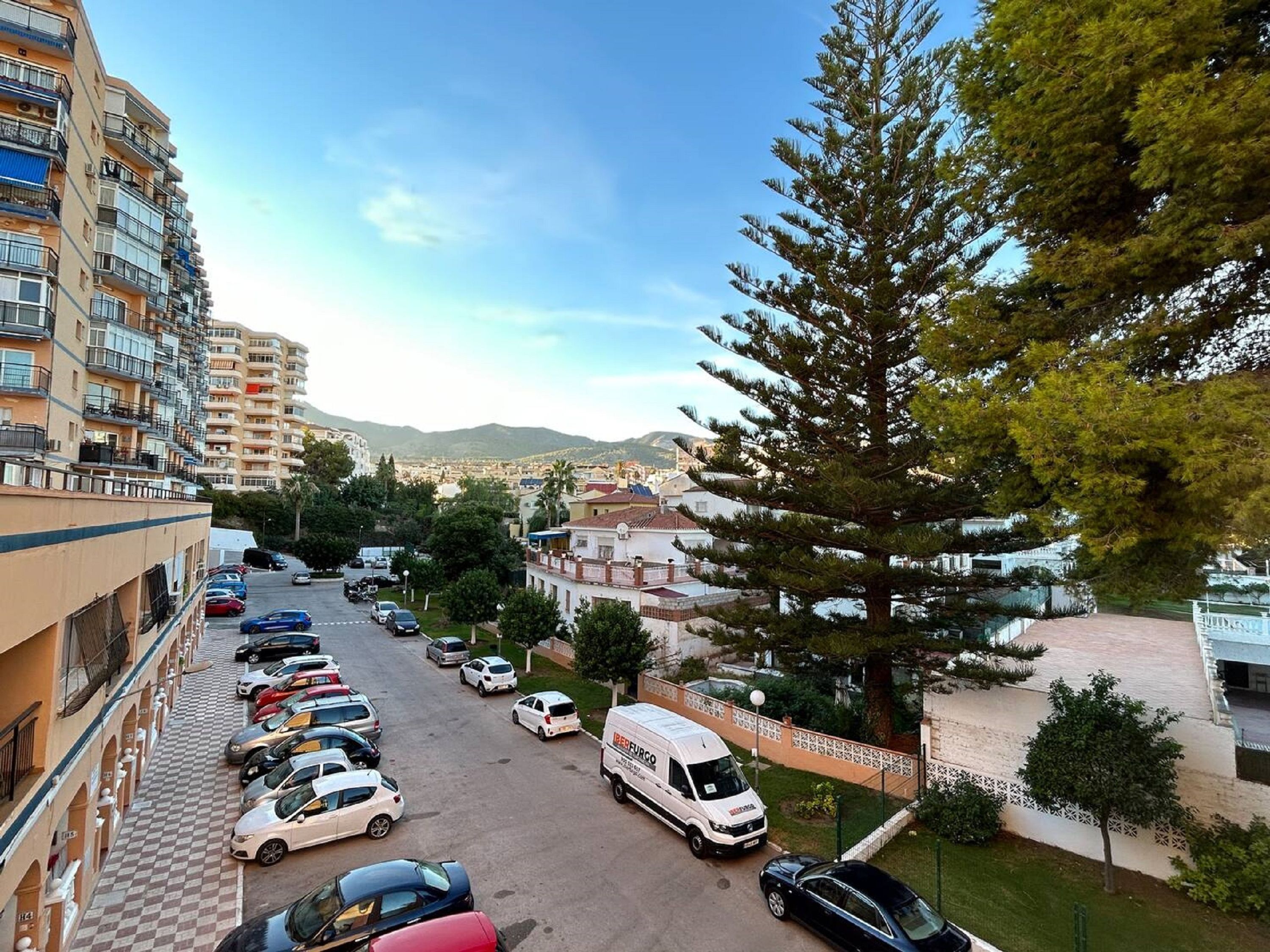 A parking lot with several cars and a delivery van, surrounded by residential buildings and a large tree.