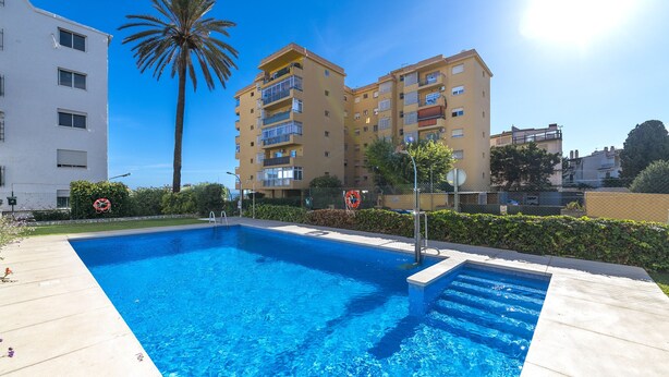 A rectangular swimming pool with a clear blue water surface, surrounded by a light-colored deck. In the background, there are two multi-story buildings with balconies and a palm tree. - Noasti de Steram Flats Torremolinos (Torremolinos)