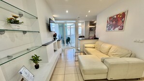 Een moderne woonkamer met een beige bank, een glazen salontafel en een flatscreen televisie aan de muur.