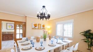 A dining room with a white table set for six, surrounded by white chairs. There is a chandelier above the table, and a large window to the right. - Real Dream Villa by Fidalsa (Jacarilla)