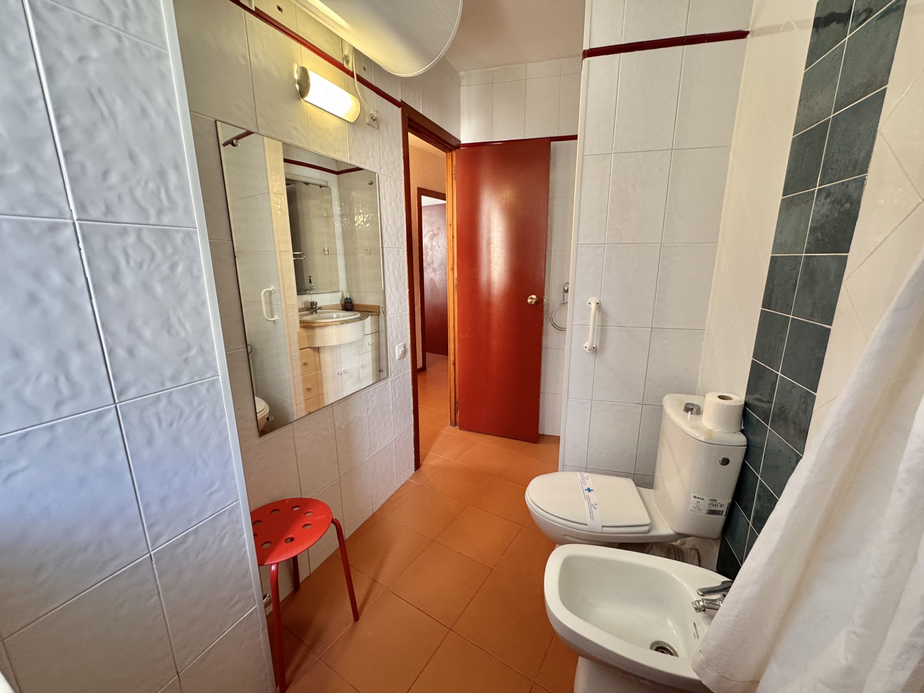 A bathroom with a toilet, sink, and mirror. There is a red chair and a door leading to another room.