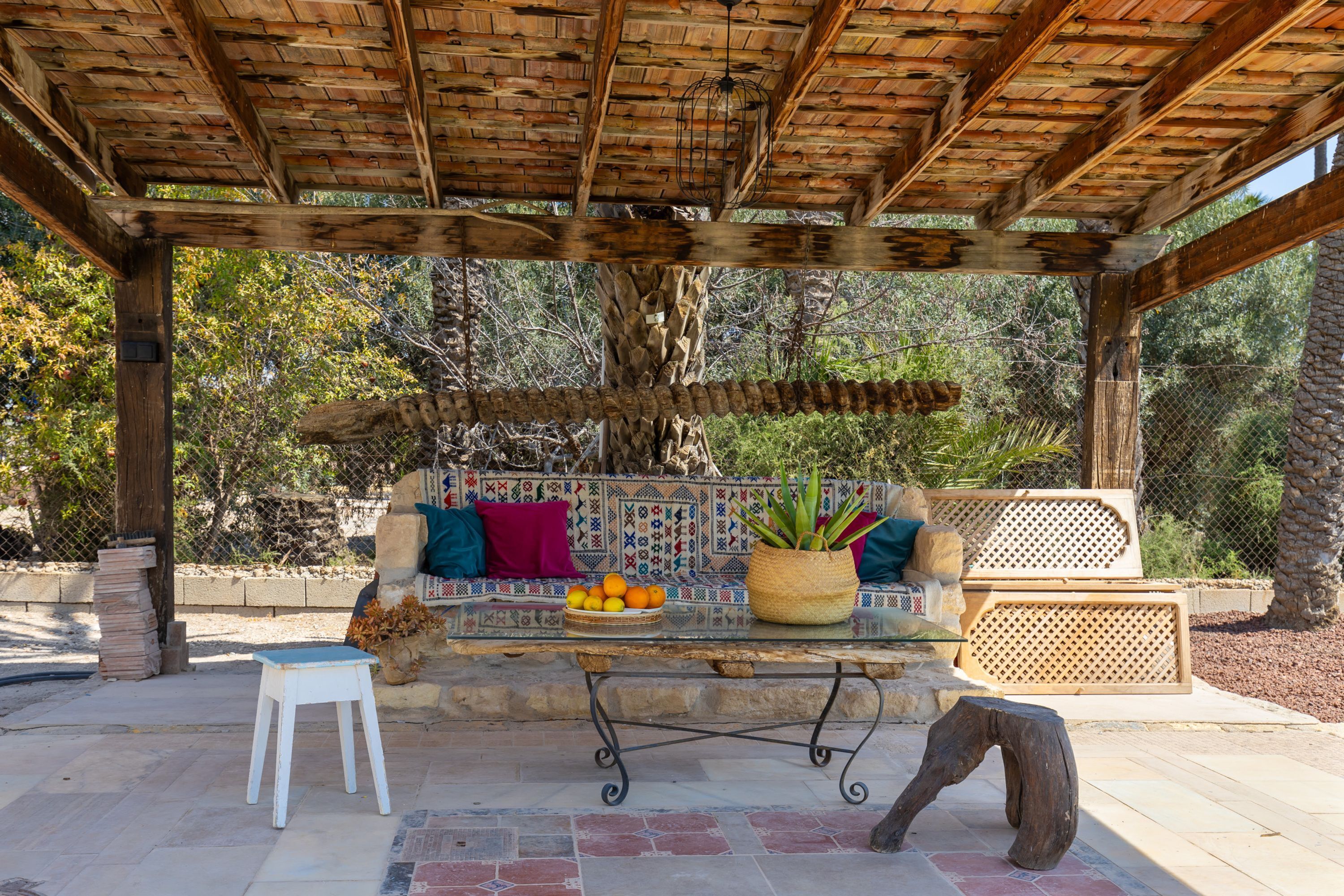 A covered outdoor seating area with a stone bench, a small table with a basket of fruit, and a white stool.
