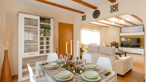 A dining area with a table set for six, surrounded by white chairs. There is a wooden cabinet with glass doors to the left, and a sofa set in the background. - Mountain Beautiful by Fidalsa (Busot)