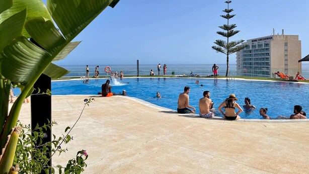A pool area with people swimming and relaxing, surrounded by tropical plants and a building labeled "ESTIVAL TORREQUEBRADA."