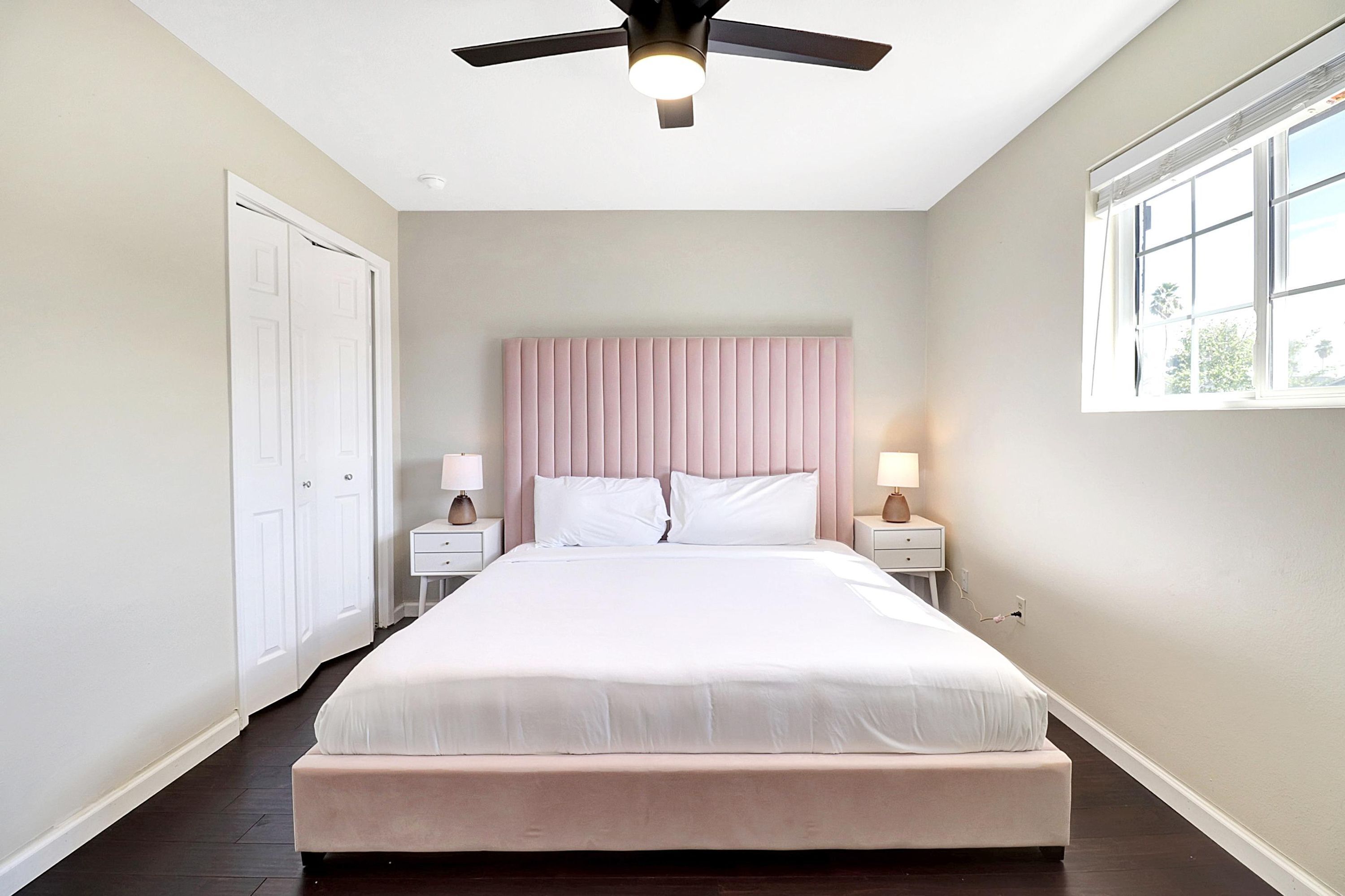 A bedroom with a large bed, a ceiling fan, bedside lamps, a window, and a door.