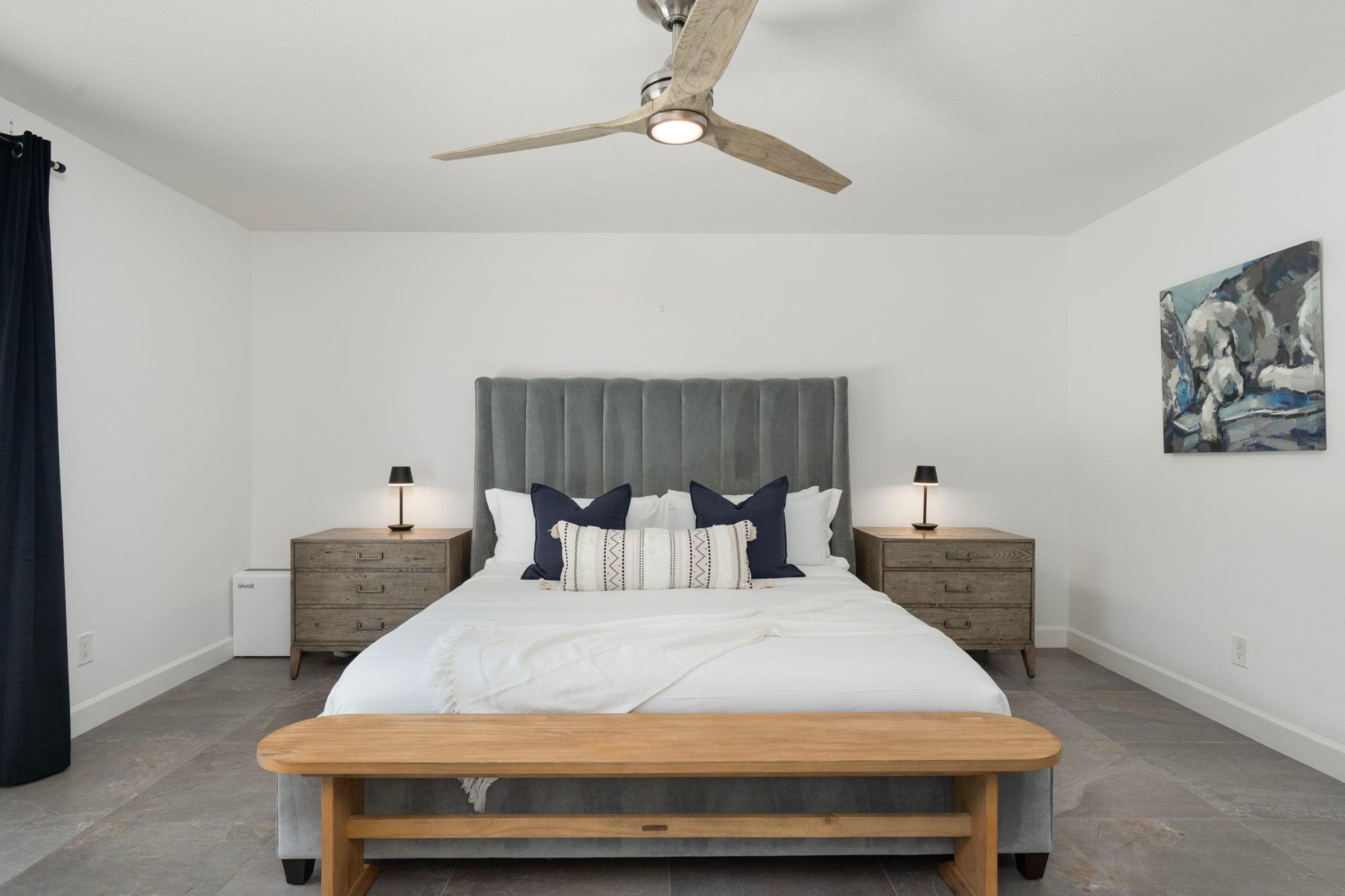 A bedroom with a bed, bedside tables, a ceiling fan, a bench, and a painting on the wall.