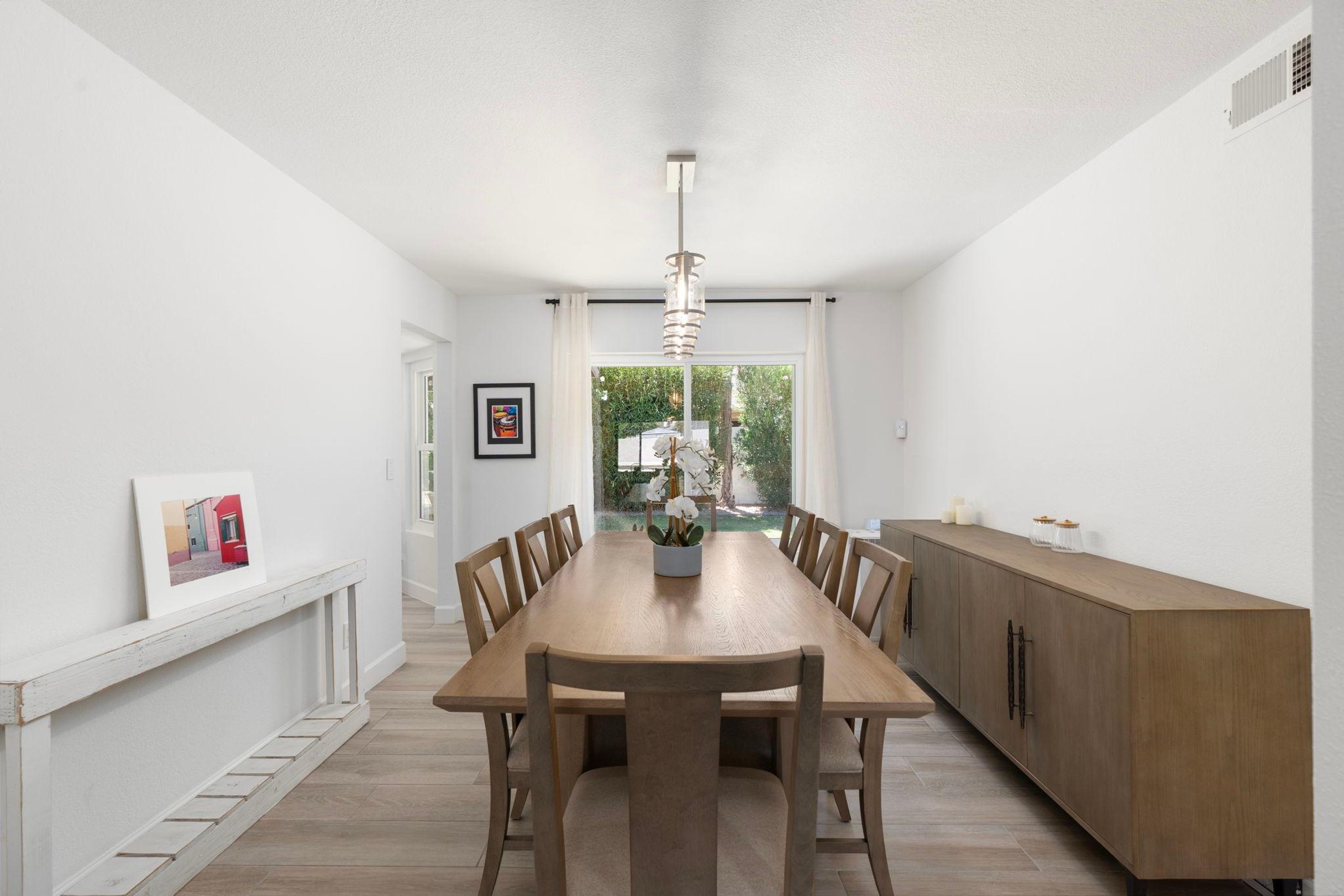 Un comedor con una larga mesa de madera, sillas, un jarrón central con flores y una lámpara colgante.