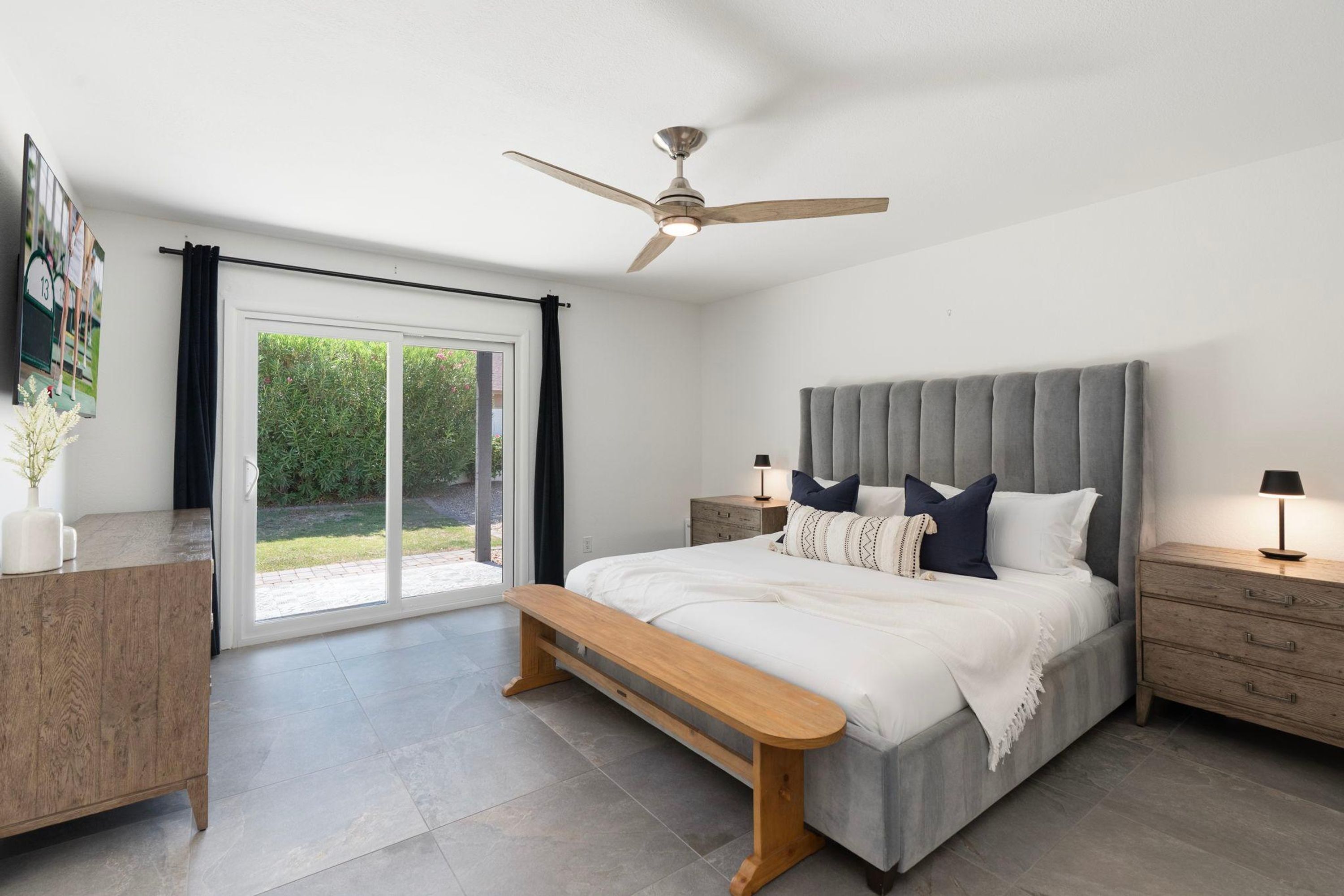 A bedroom with a large bed, a ceiling fan, a bench, a nightstand, and a sliding glass door leading to a garden.