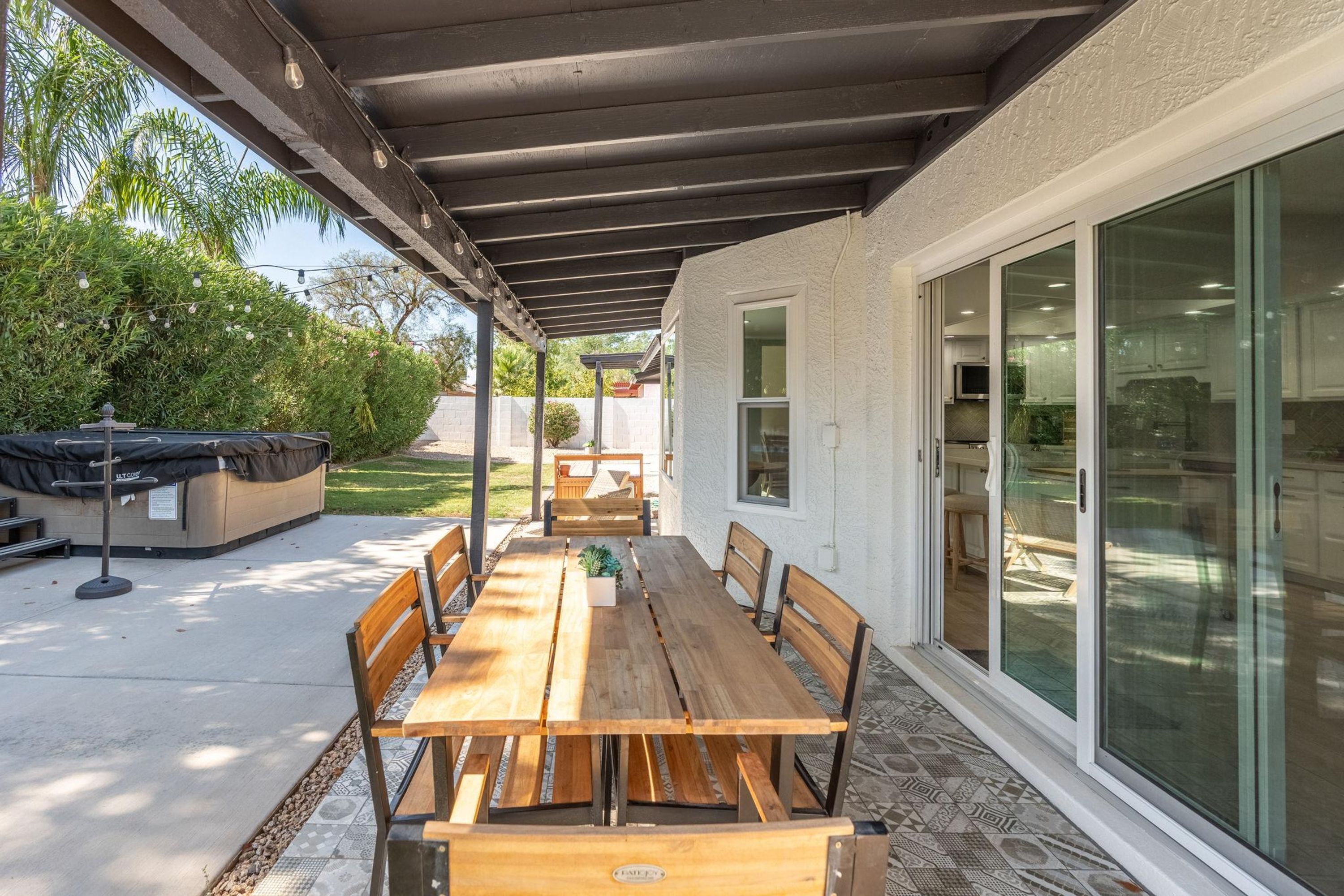 An outdoor patio with a wooden table and benches, a covered area with string lights, and a view of a house with sliding glass doors.