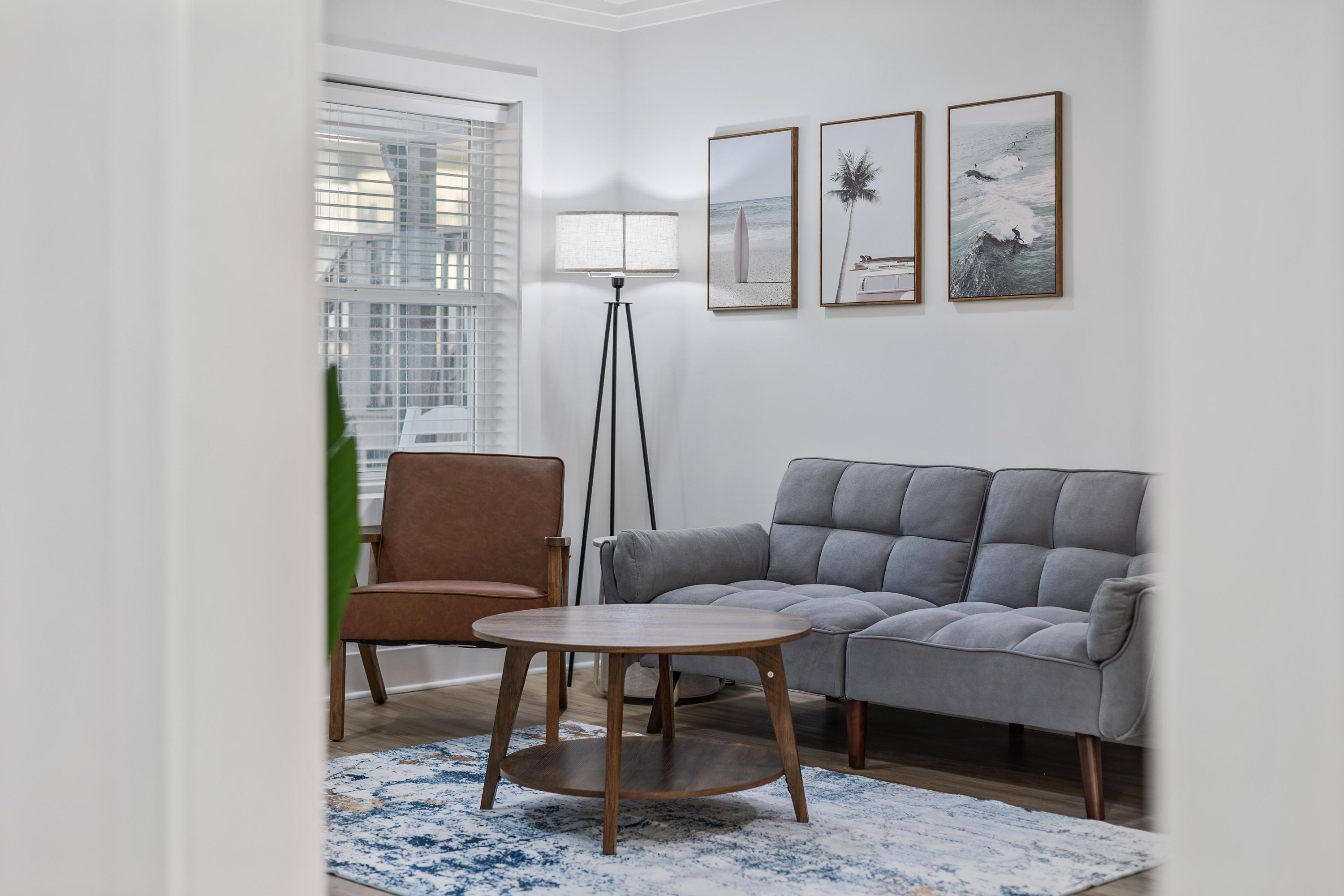 Una sala de estar con un sofá gris, una silla marrón, una mesa de centro redonda de madera y una lámpara de pie. Hay tres cuadros enmarcados en la pared.