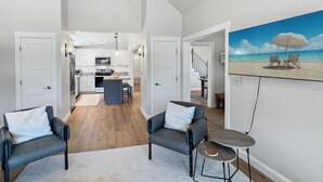 A modern living room with a kitchen in the background, a painting of a beach scene, and a staircase leading to another level. - Long Sands Your All-season Getaway (York)